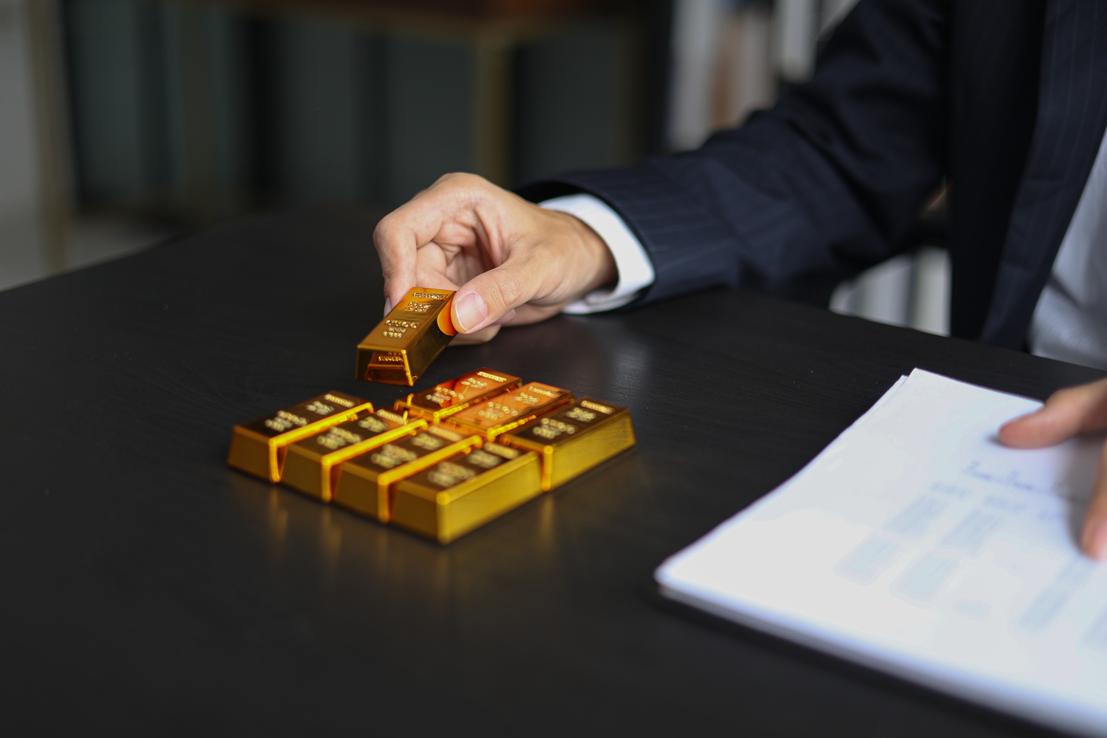In Edelmetalle investieren – einfach bestellt, sicher gekauft Das Bild zeigt eine Hand, die auf einem Tisch Goldbarren stapelt.